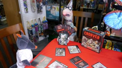 A plush wolf in a white coat and a strange doctor-like figure with a very large nose sit at a table playing a card game. The cards have images of comical corpses and spooky items on them.  