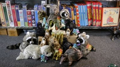 A cardboard box can be seen peeking out from under a plethora of puppets of various sizes, including raccoons, crows, a large donkey, dragons, a funny mouse-like creature, and some mini frogs.