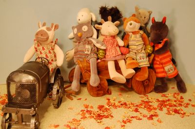 A plush cow in a dress drives a tractor pulling a wooden wagon overflowing with animals wearing clothing.