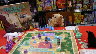 A plush capybara, axolotl and little crow are seated at a table with a colorful gameboard. The pieces, of various colors, are of pyramids in different heights of completion.