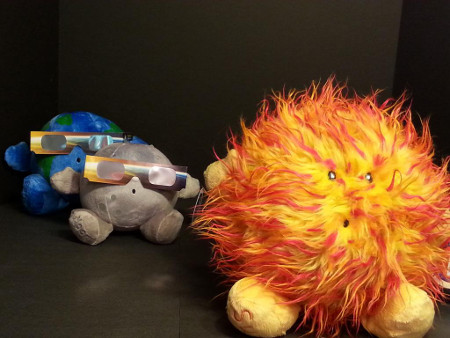 A plush Earth sits behind a plush moon; both are wearing eclipse sun glasses. In front is a big, plush sun.